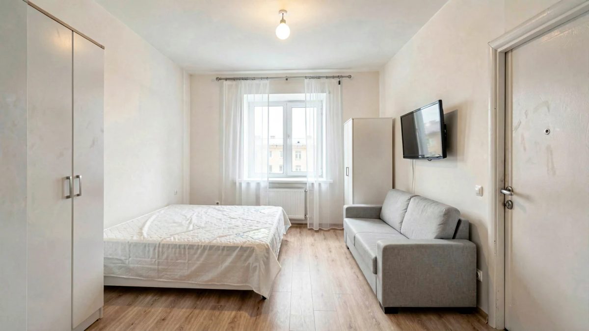 Однушка в хрущёвке после ремонта: комната около 18 м², кровать у левой стены, серый диван и телевизор напротив, белые шкафы у входа