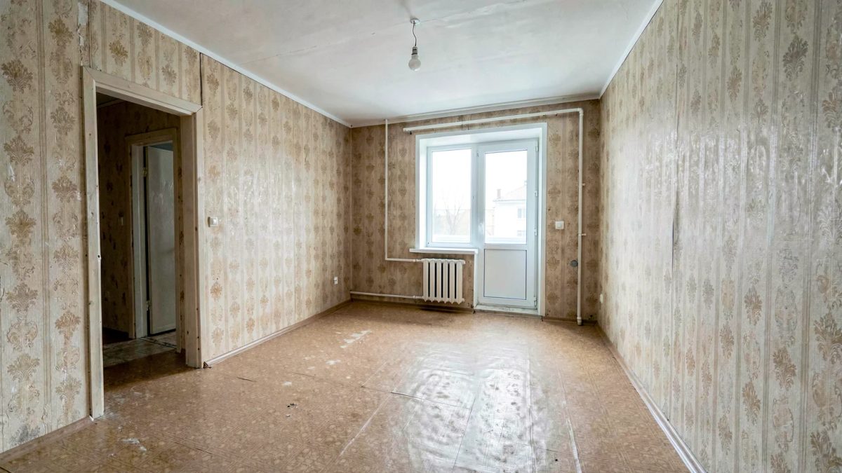 Комната однушки 18 м² до ремонта: выцветшие обои, линолеум, лампочка на проводе, окно и балконная дверь в торце, проём в прихожую слева